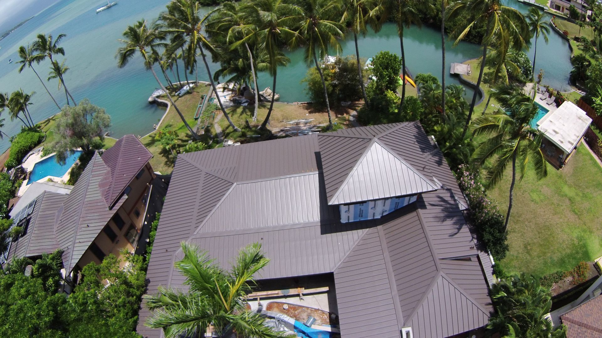 Aerial view of a brown-roofed house with a swimming pool, near a waterway with palm trees and boats.