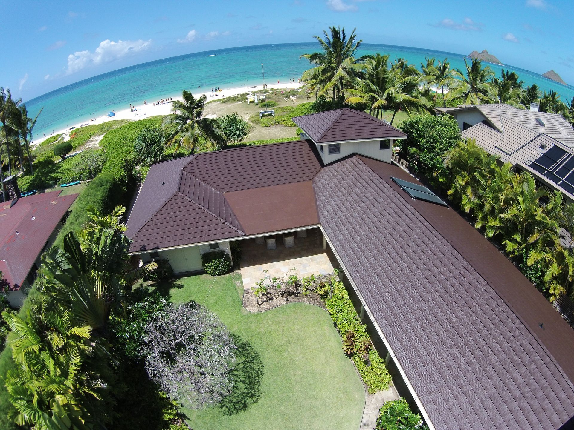 Aerial view of a brown-roofed house with green lawn near a beach with white sand and turquoise water.