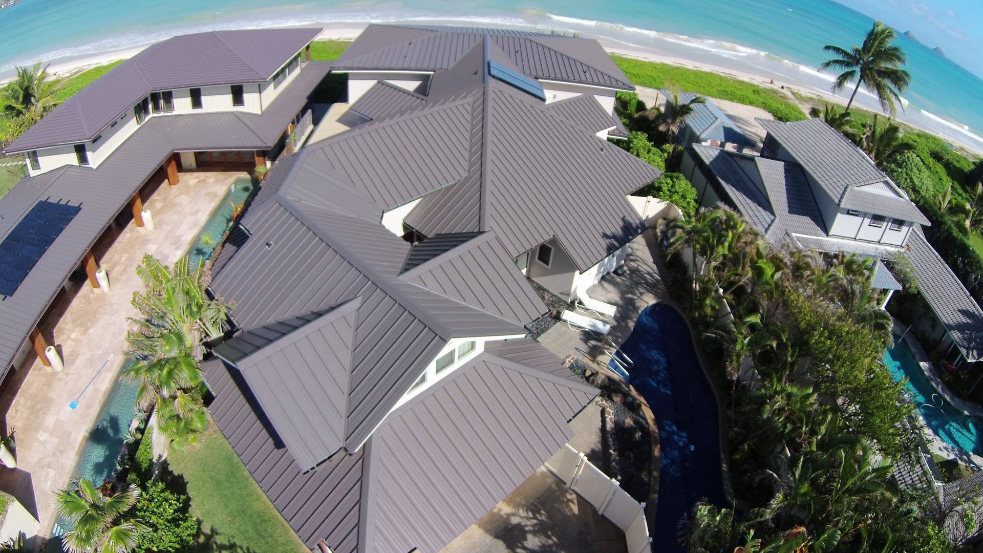 Aerial view of large, multi-sectioned house with dark roof next to a beach and ocean.