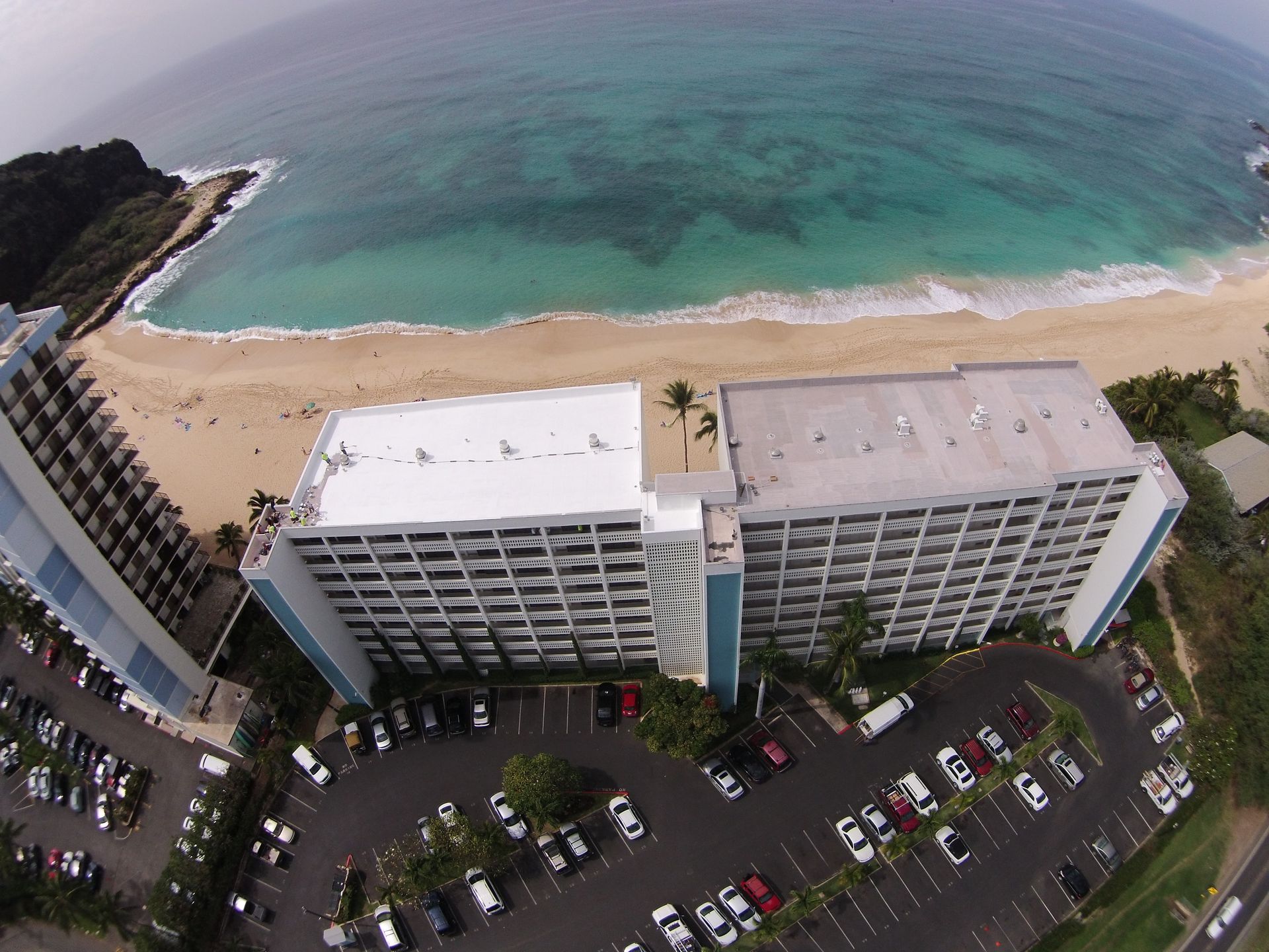 Aerial view of a beachfront hotel and parking lot, sandy beach, and turquoise ocean.
