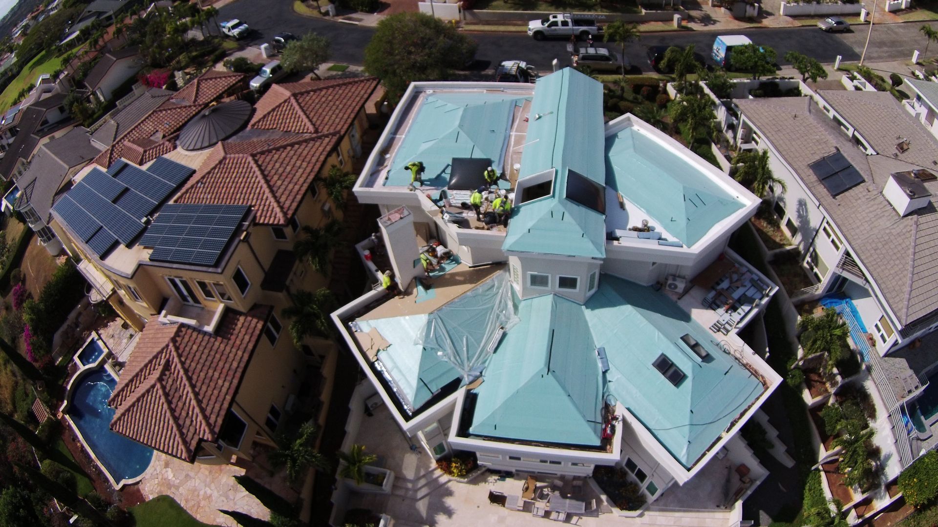 Aerial view of a home with turquoise roof and workers, surrounded by other houses with brown tile roofs.