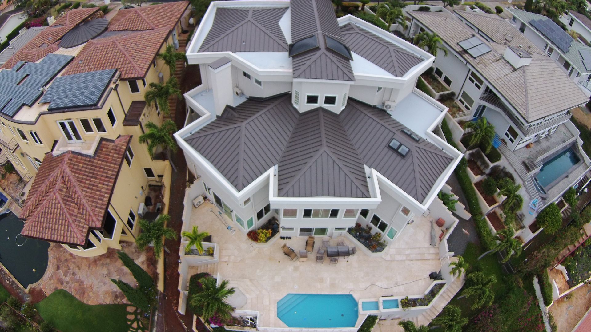 Aerial view of a large white house with a dark gray geometric roof, a swimming pool, and surrounding houses.