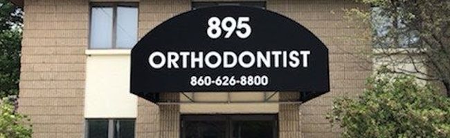 Building entrance with a black awning displaying 895 ORTHODONTIST and a phone number in white text, surrounded by beige brick and greenery.