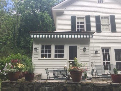White house with black shutters and a green-striped awning. A stone patio features potted plants and an outdoor dining set, surrounded by trees. Calm and inviting.