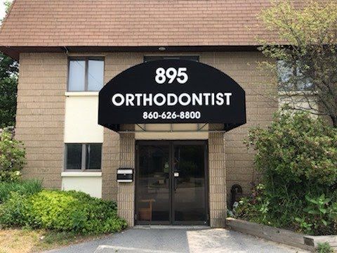 Exterior of a brick orthodontist office with a peaked roof. A black awning displays the address and phone number. Surrounding greenery adds a welcoming feel.