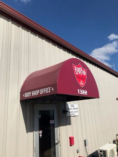 Red awning covers a door on a beige building. A shield logo is visible. Bright, clear blue sky.