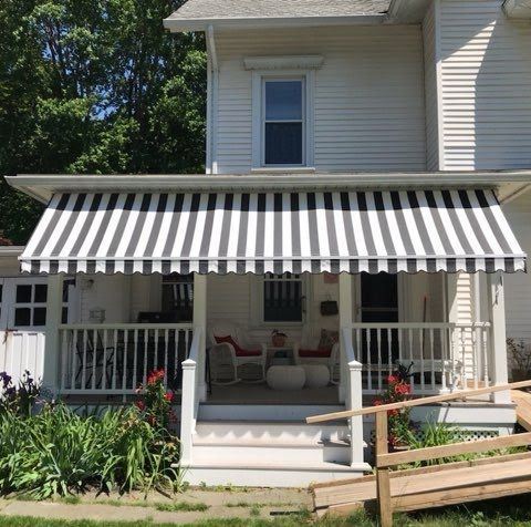 White house with a covered porch featuring black-and-white striped awning. Red chairs on porch and vibrant garden. Sunny and welcoming atmosphere.