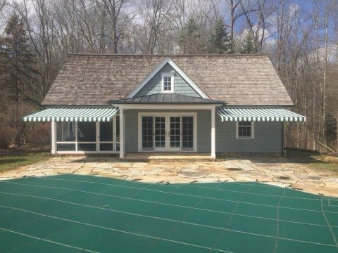 A quaint cottage with a steep shingle roof stands against a backdrop of bare trees. Striped green awnings shade the windows. A closed pool lies in the foreground.