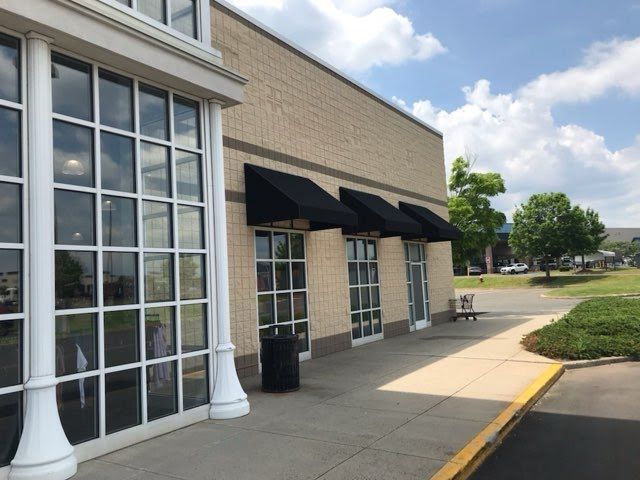 Outside a building with large windows and black awnings, a sunny sidewalk leads to a parking area with greenery. The sky is partly cloudy, creating a calm ambiance.
