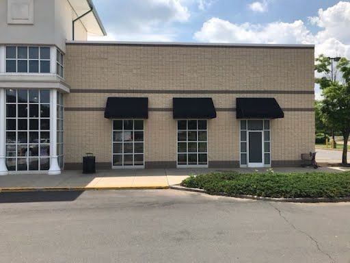 A beige brick storefront with three large windows, each topped by black awnings. Green bushes and a clear sky add to the calm, suburban scene.