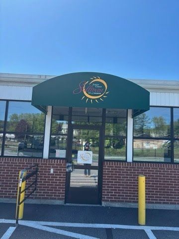 A brick building entrance with a glass door and a dark green awning displaying a logo. A sunny day with trees and a clear blue sky in the reflection.