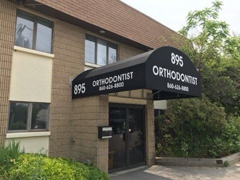 Two-story building with a brown brick facade, featuring an entrance canopy. Sparse greenery surrounds the path.