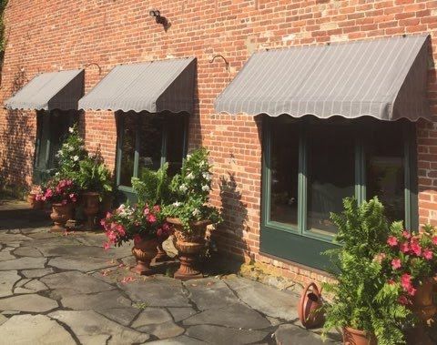 Brick wall with three windows shaded by gray awnings. Flower pots with lush green plants and pink flowers line the stone-paved path, creating a charming, serene atmosphere.
