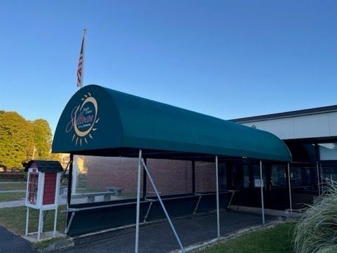 A green awning entrance with a logo leads into a building under a clear blue sky. An American flag waves in the background.