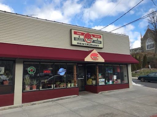 Storefront of a restaurant with beige siding and a red awning, displaying signs for pizza and salads. Potted plants and neon signs are visible in the window.