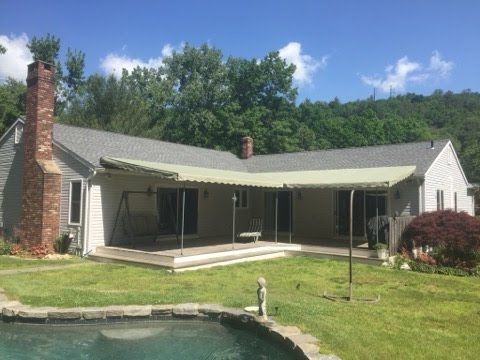 A single-story house with a covered patio, brick chimney, and sliding doors. The yard features green grass, a small pool, and lush trees in the background.