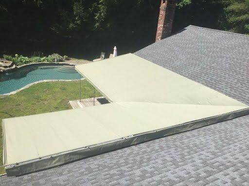 A green awning covers a gray shingled roof, casting shade over a backyard. A pool with curved edges and lush greenery is visible in the sunny background.
