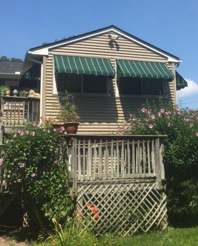 Two-story house with beige siding, green-striped awnings, and a wooden deck. Surrounded by blooming pink flowers and lush greenery under a clear blue sky.