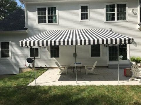 A backyard patio features a black-and-white striped awning providing shade over a seating area with white chairs and a table, against a white house facade.