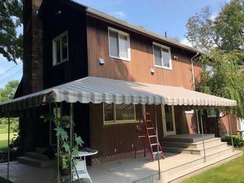 A two-story brown house with white awning shades a patio. A red ladder leans against the house, surrounded by green trees on a sunny day.