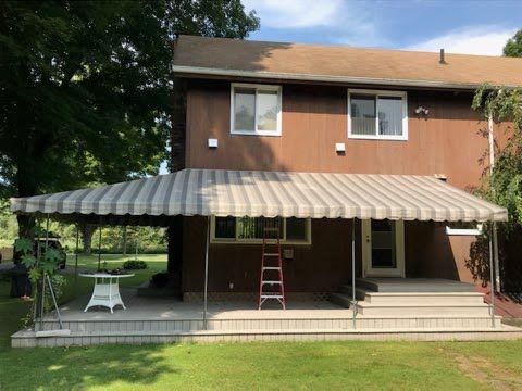 Two-story brown house with a striped awning over a back patio. A red ladder is on the porch, with a white table and greenery around. Sunny atmosphere.