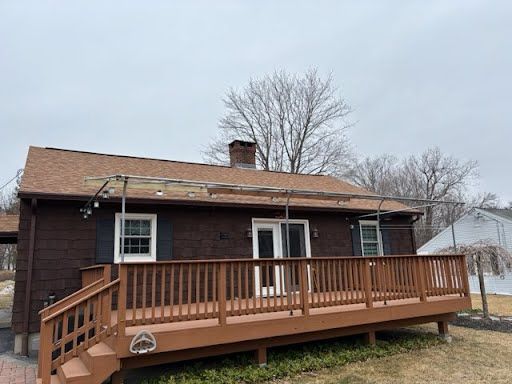 A small brown house with a wooden deck and railing. Overcast sky, barren trees in the background, and string lights above the deck add a quaint feel.