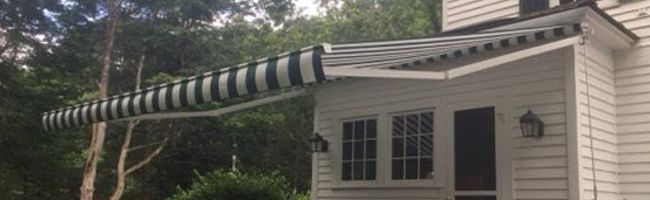 A white house with a green and white striped retractable awning extends over a porch. Lush trees surround the house, creating a serene atmosphere.