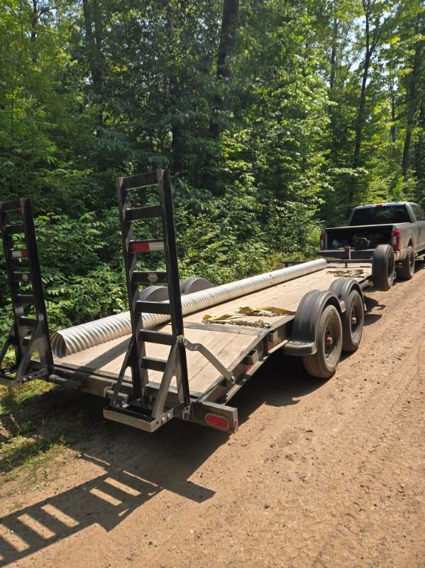 Utility trailer carrying long white pipes on a dirt road beside a wooded area.