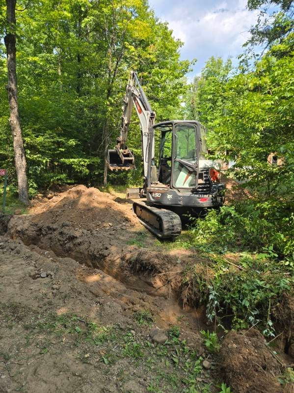 Excavator digging a trench in a wooded area with fresh soil and green trees nearby.