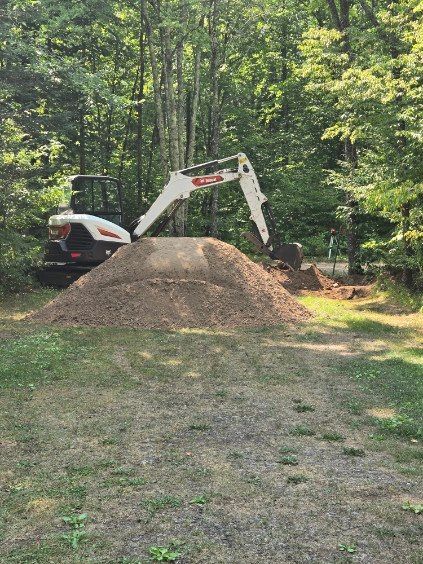 Small excavator beside a dirt mound in a wooded yard.
