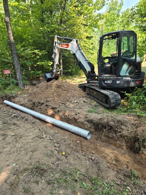 Small excavator digging a trench in a wooded area beside a large metal pipe.