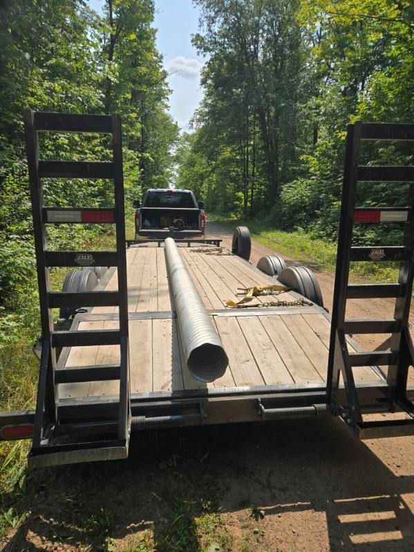 Flatbed trailer with metal ramps, a large pipe, and wooden planks in a wooded area.
