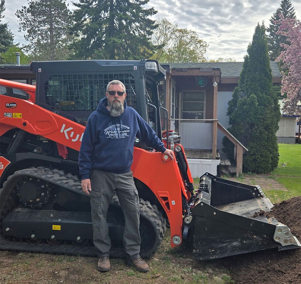 Man standing beside an orange skid steer loader with a bucket in a yard by a house
