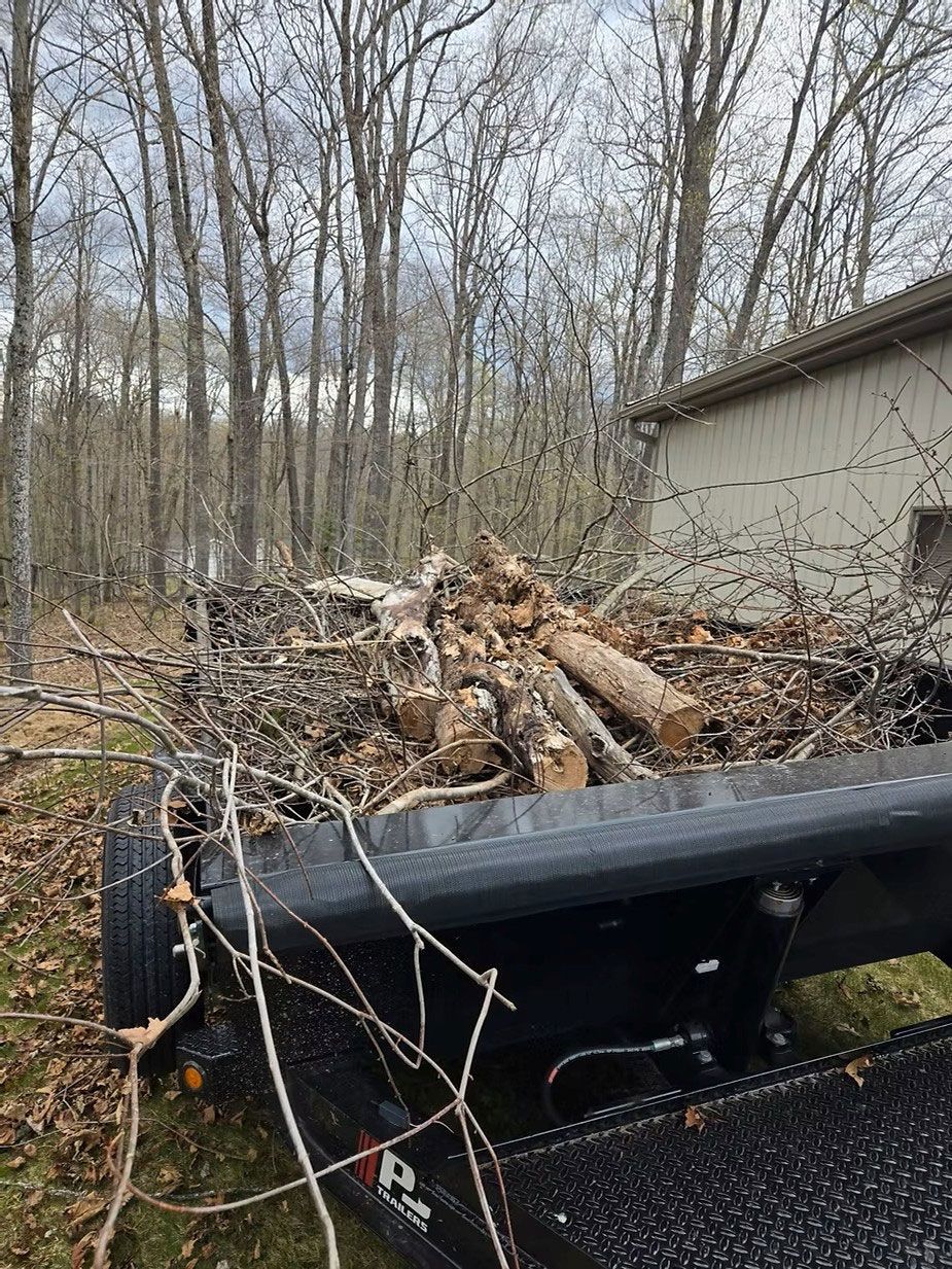 Flatbed trailer loaded with branches and logs beside a house in a wooded yard.