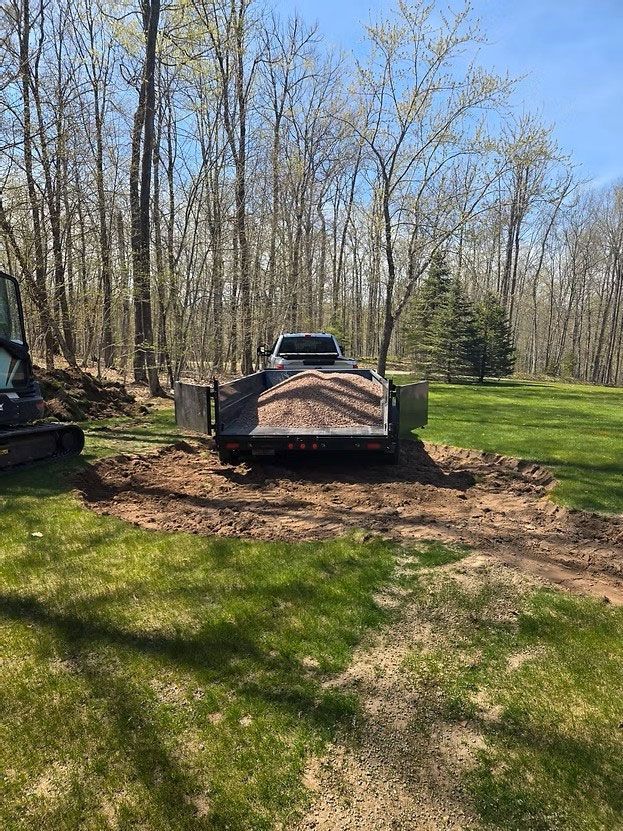 Truck hauling dirt on a trailer in a wooded yard beside grass and bare trees.