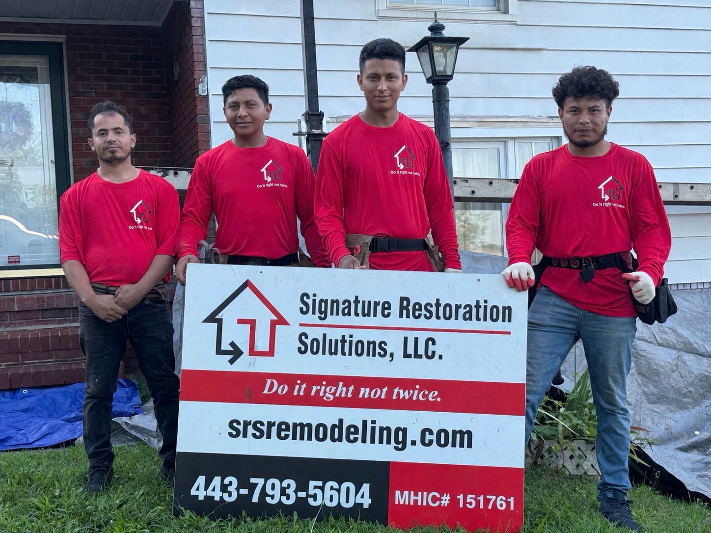 Four people in matching red shirts stand outside a house holding a Signage Restoration Solutions, LLC business sign.