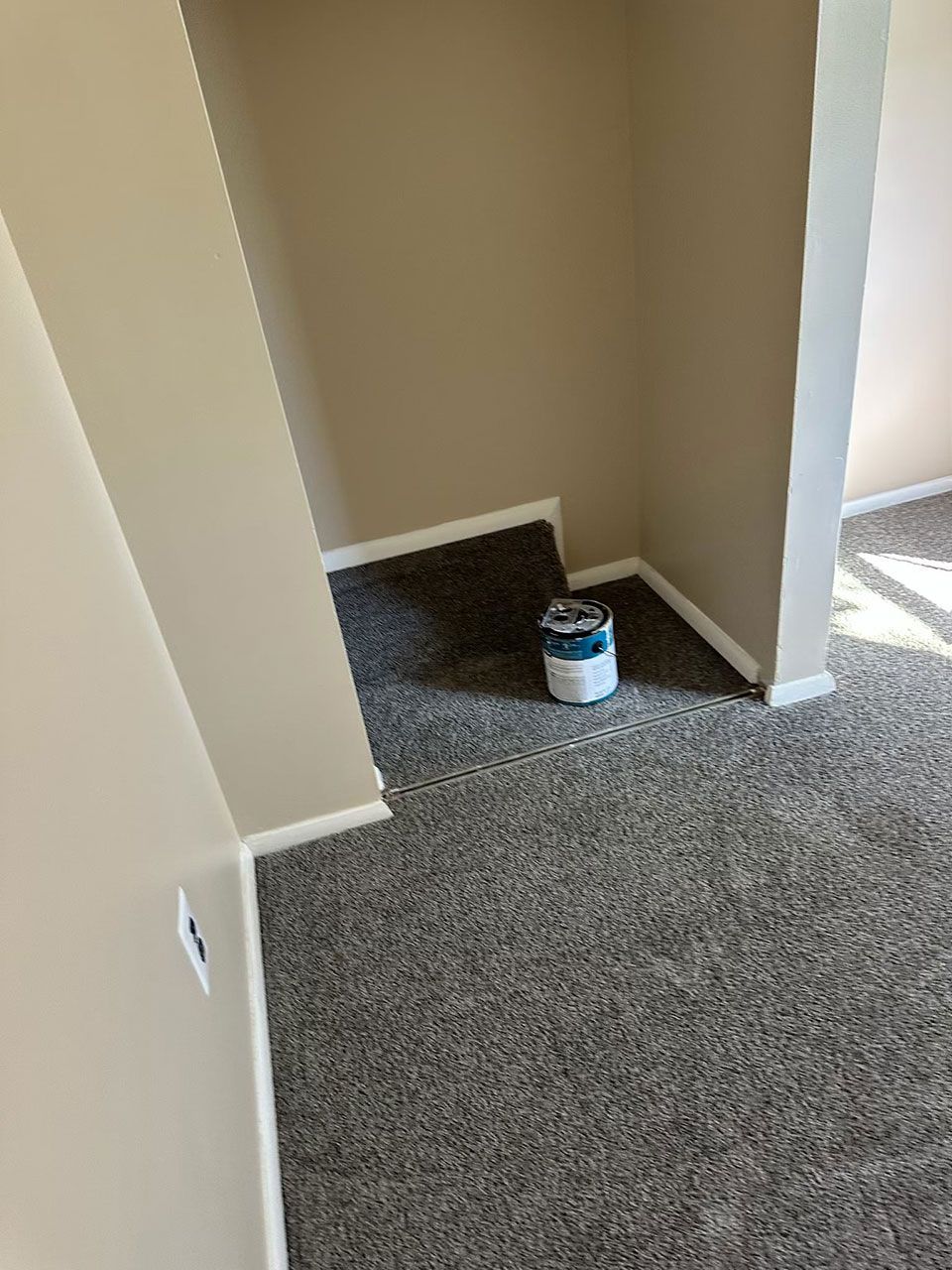 A can of paint sits in a room with new gray carpet and beige walls.