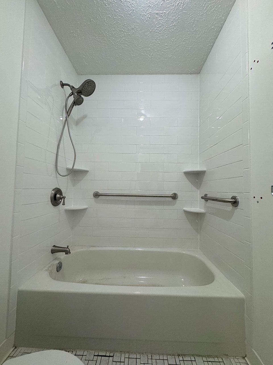 White bathroom with bathtub, shower head, grab bars, and shelves.