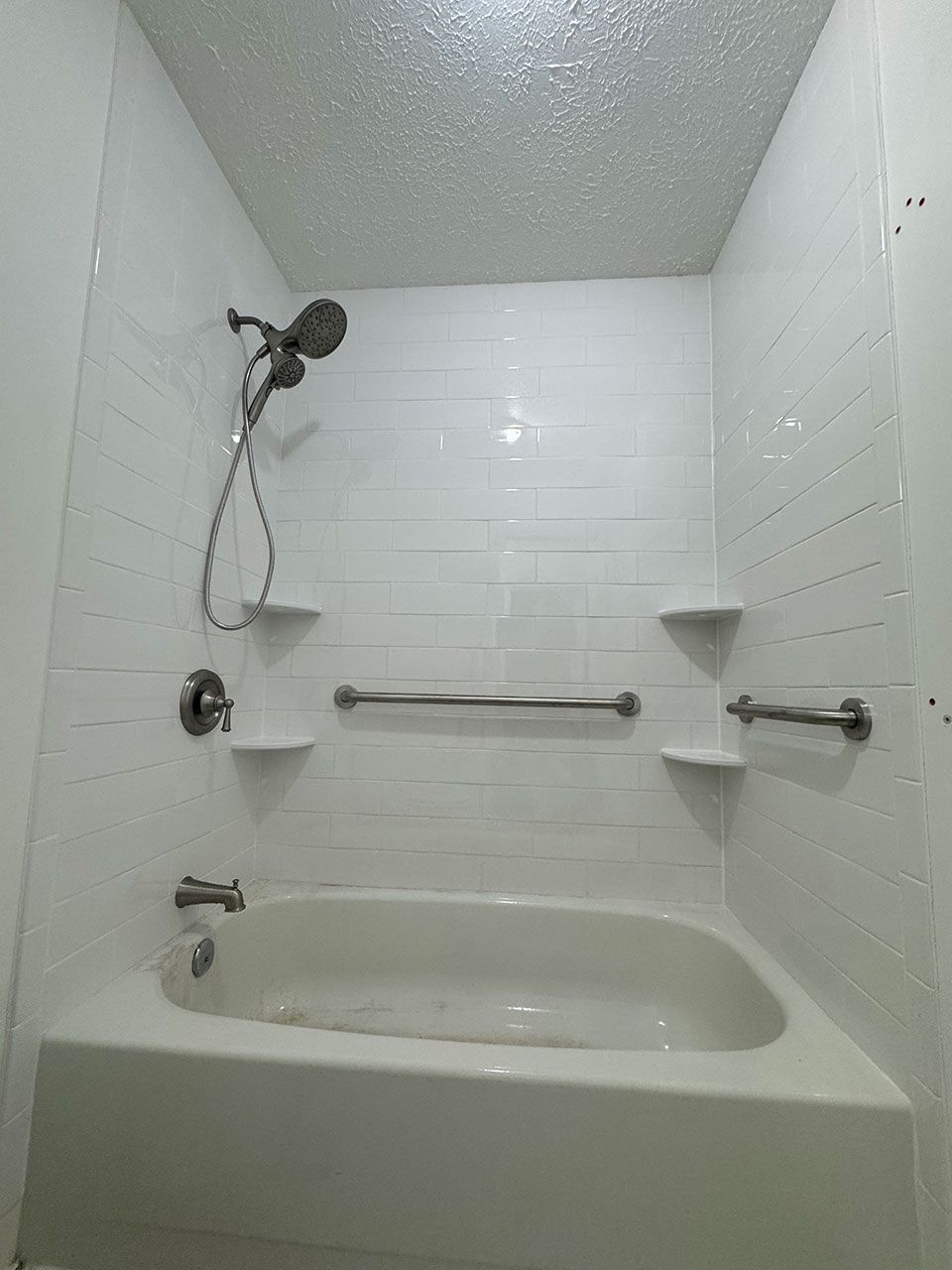 White bathtub with shower, horizontal tile, handrails, and built-in shelves.