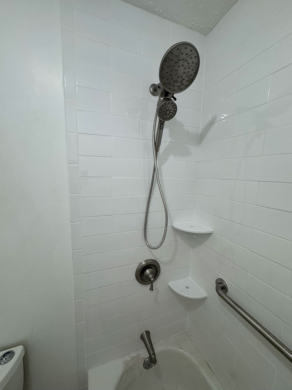 White tiled shower with handheld shower head, grab bar, and two shelves.