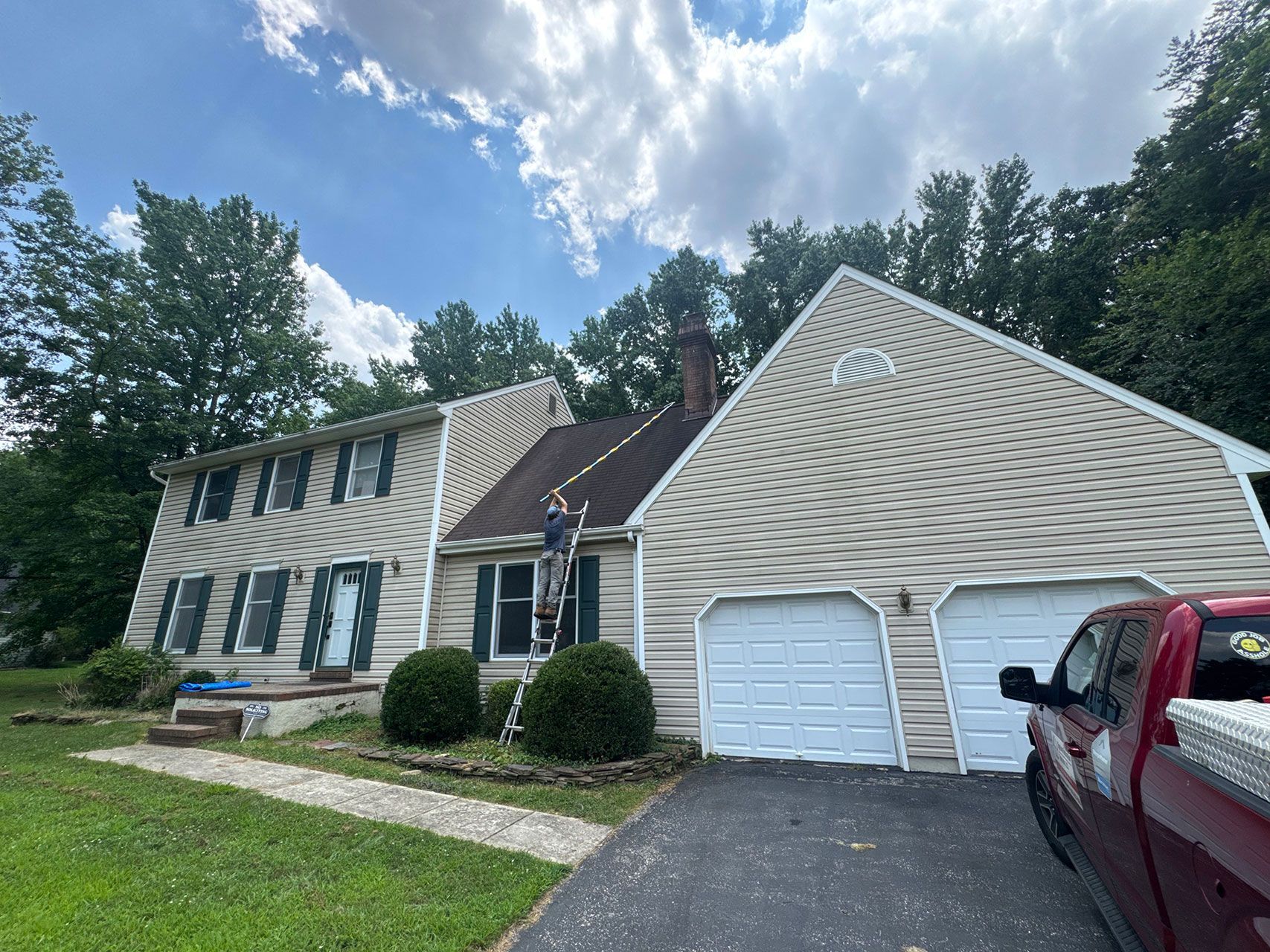 Two-story house with tan siding, a red truck, and a person on a ladder on the roof.