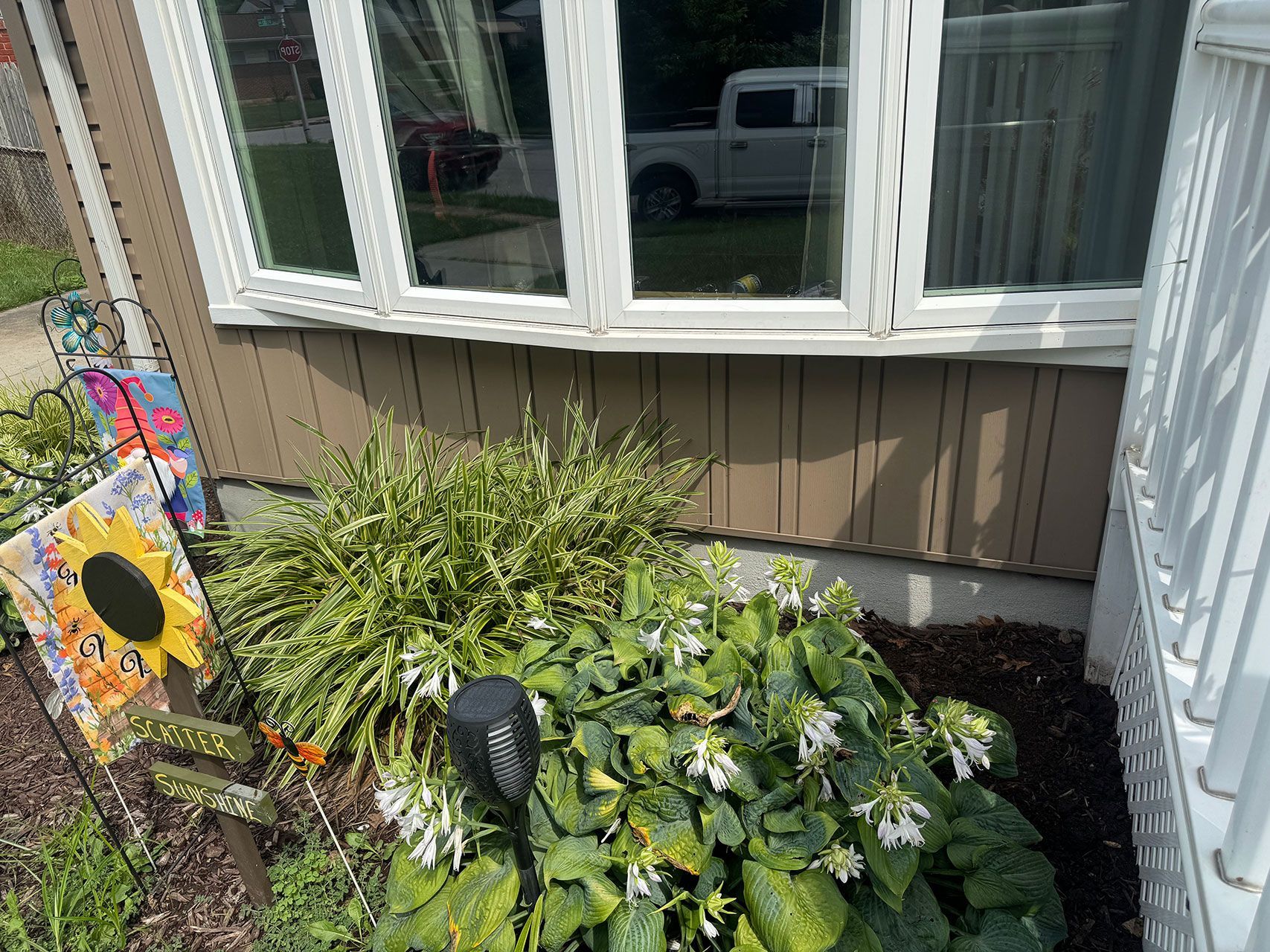 Close-up of a house with brown siding, a window, and a garden with plants and a sunflower decoration.