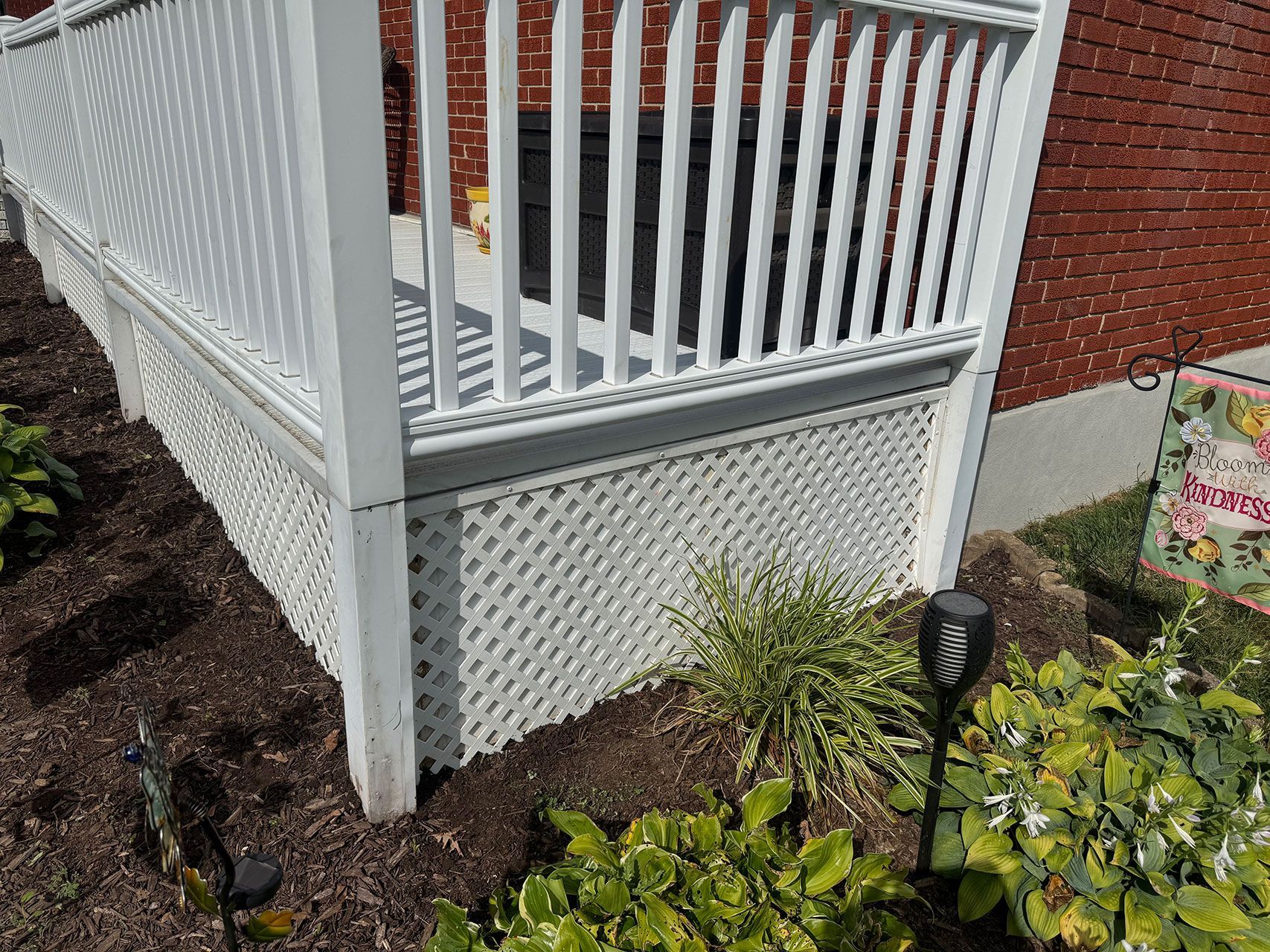 White lattice skirting under a deck, with white railing. A solar light and green plants are in the flower bed.