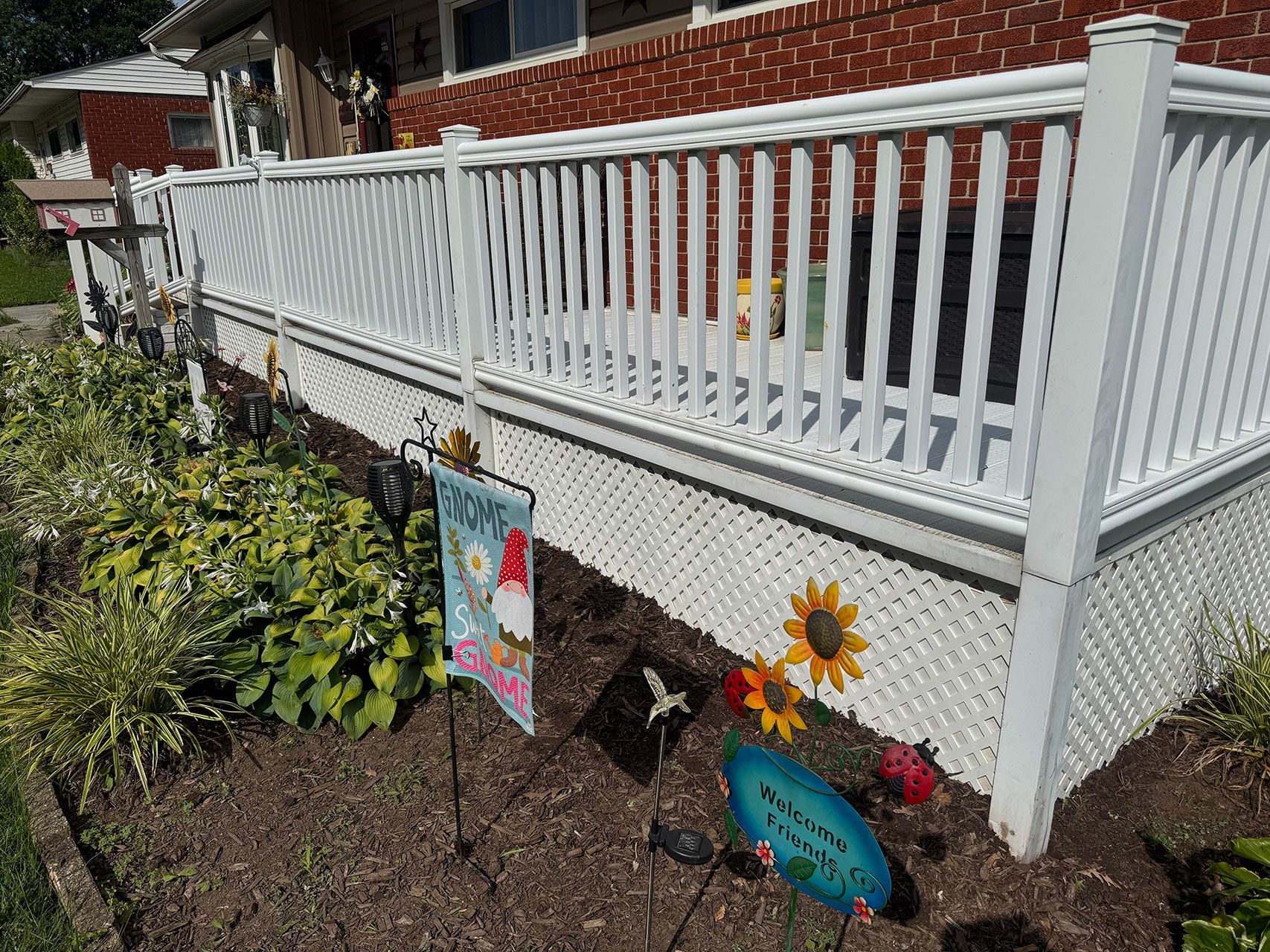 White railing on a brick house with a flower bed in front, decorated with garden signs and metal flowers.