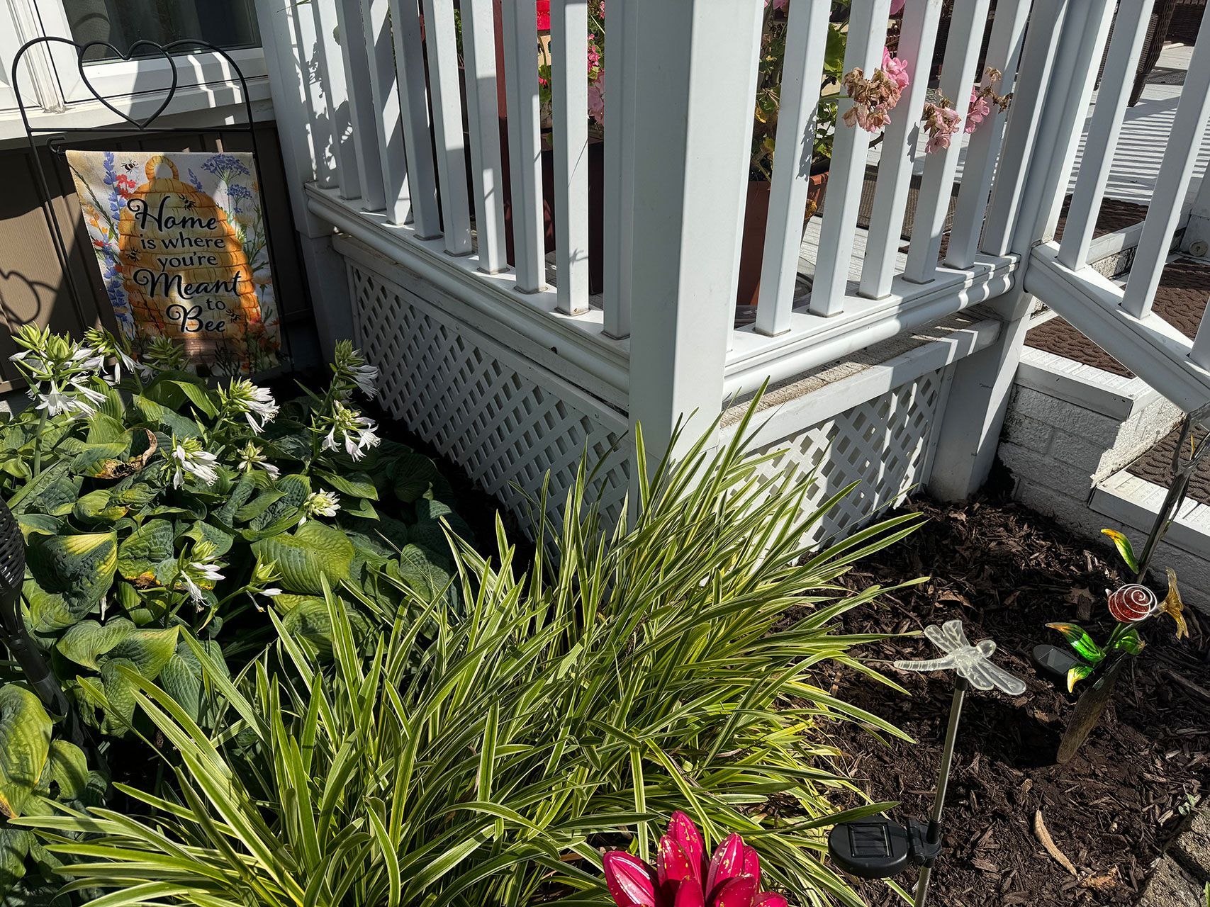 Garden bed with white railing, sign, and various plants, including green and yellow grasses and pink flowers.