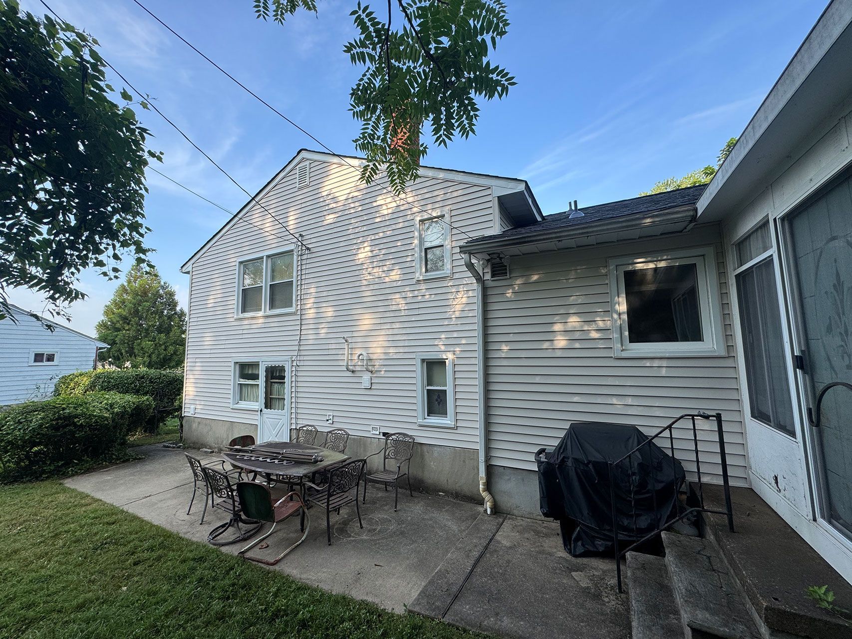 Back of a two-story white house with a patio, table, chairs, grill, and a side structure.