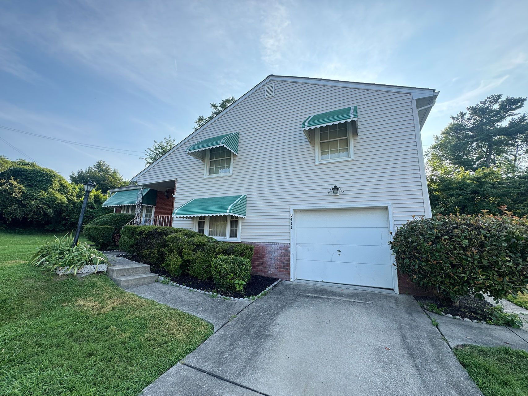 Two-story house with green awnings over windows, a white garage door, and a concrete driveway.
