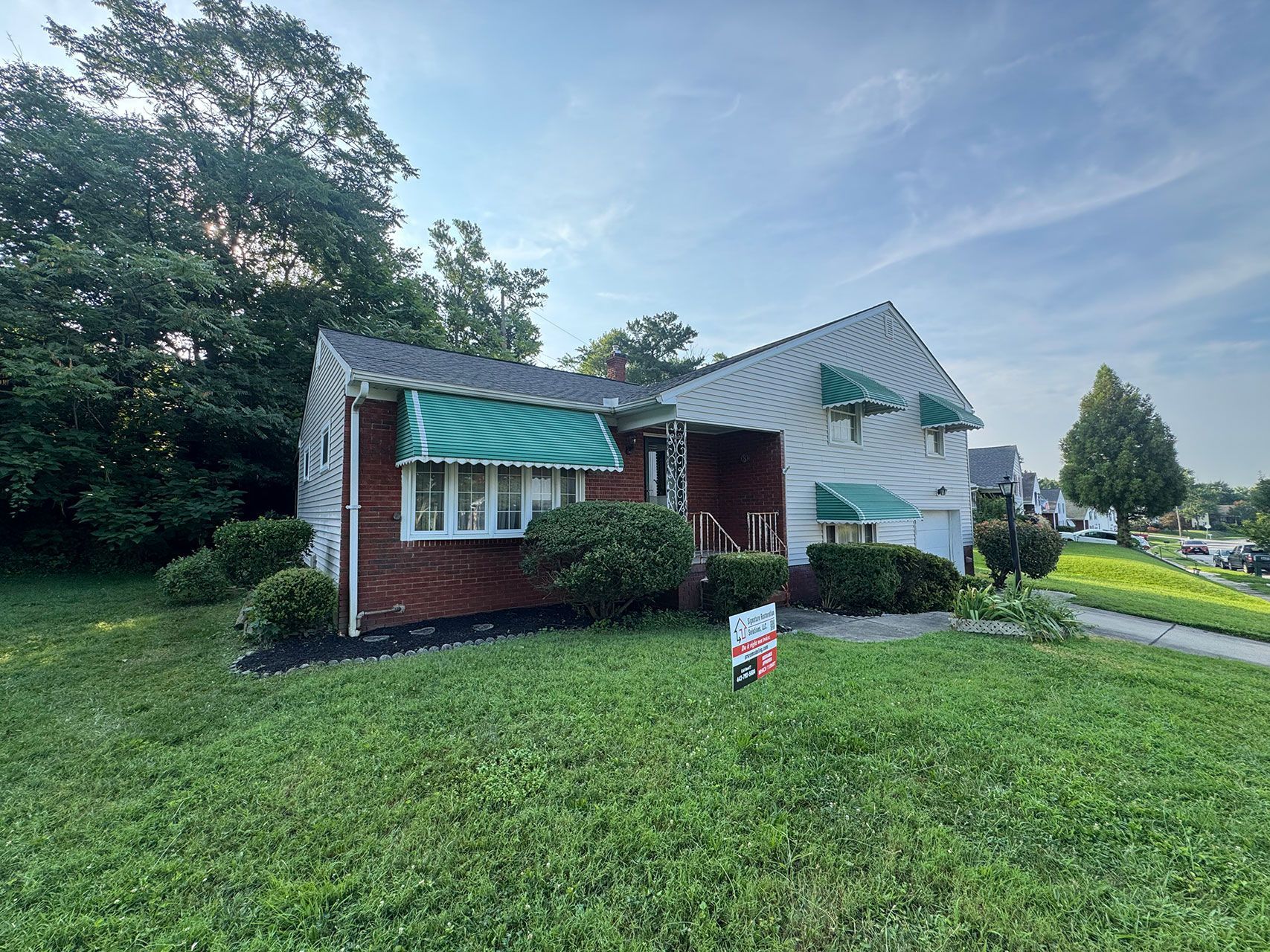 A two-story house with a red brick facade, green awnings, and a for sale sign on the front lawn.