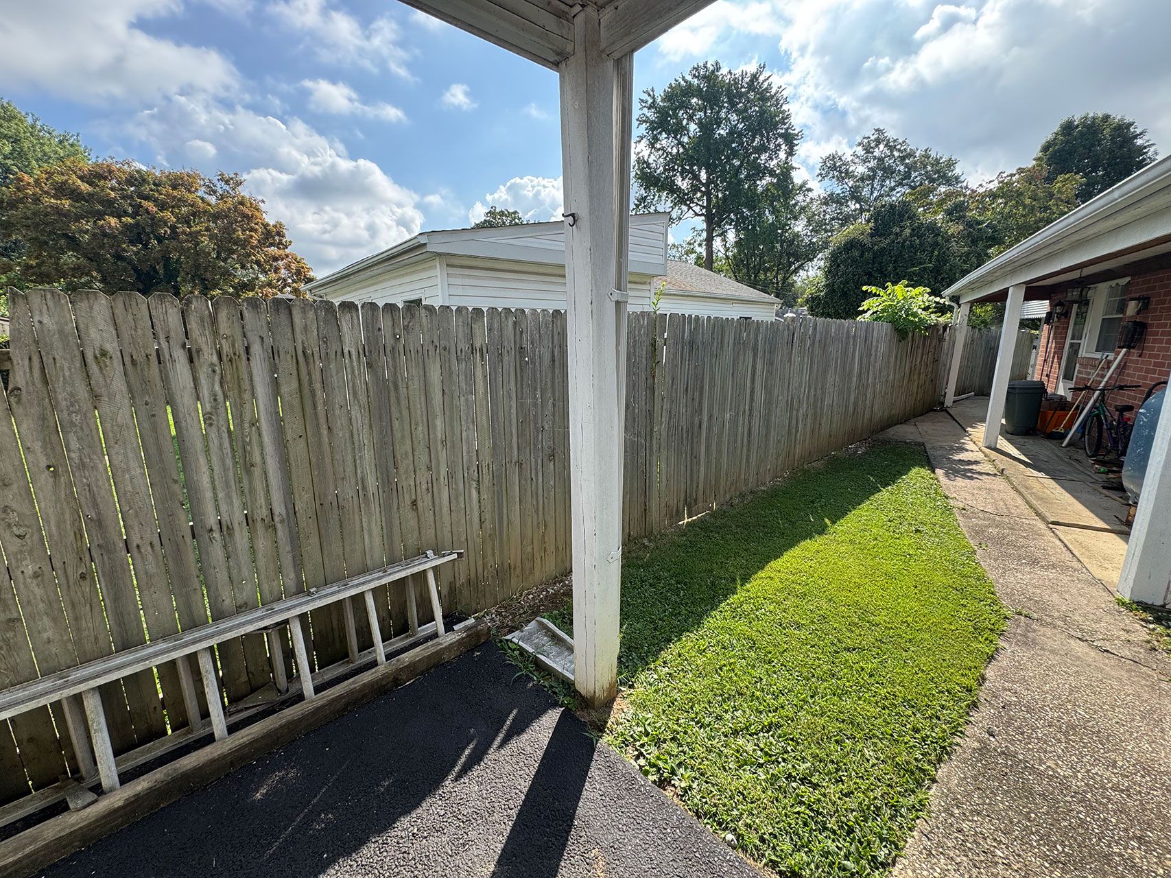 A wooden fence, small green lawn, and walkway.  A white pillar supports a roof. Sunlight casts shadows.
