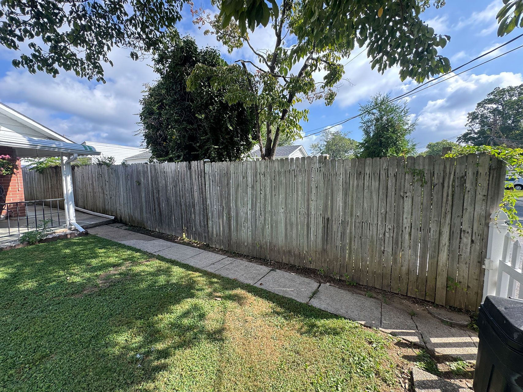 Wooden fence along a concrete path in a backyard, under a cloudy sky. Green grass, trees.